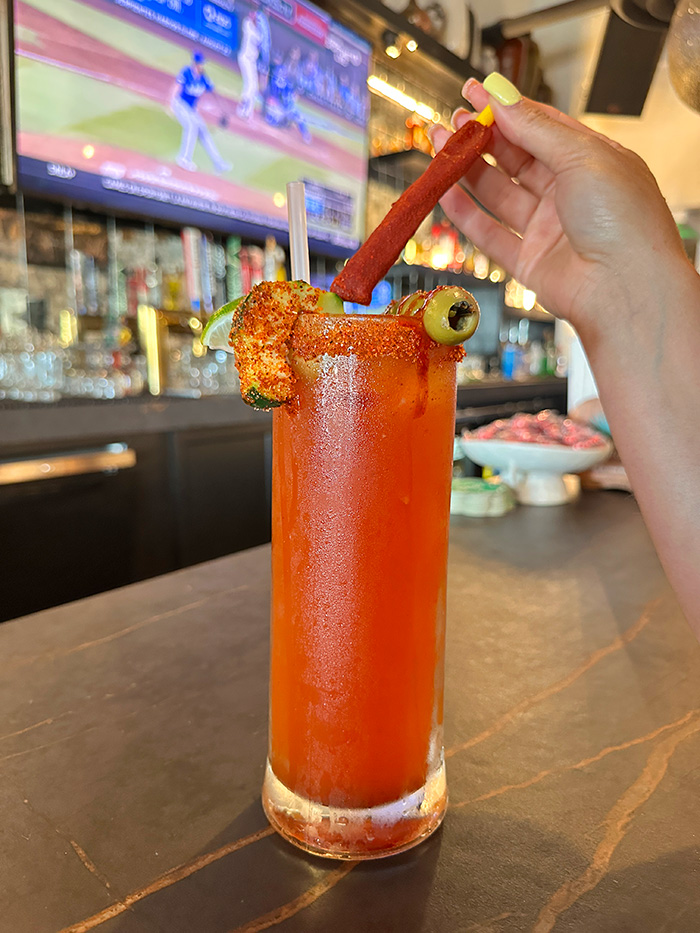 Tall red michelada with tajin rim, olive and tamarindo straw held over a bar