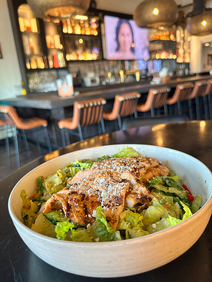 Chicken Caesar salad in a white bowl with the bar and leather stools blurred behind