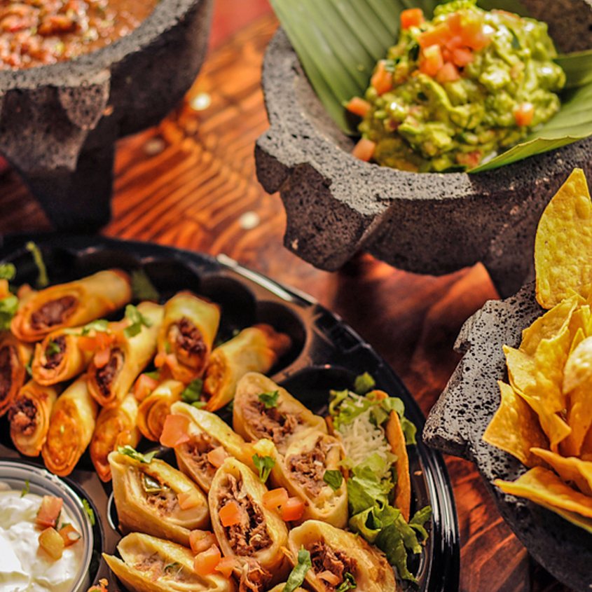 Party platter close-up with taquitos, mini quesadillas, chips, salsa, and guacamole in stone molcajetes on a wooden table