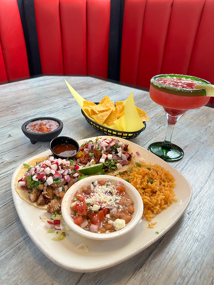 Three street tacos with radishes and cilantro alongside charro beans, chips and a pink margarita